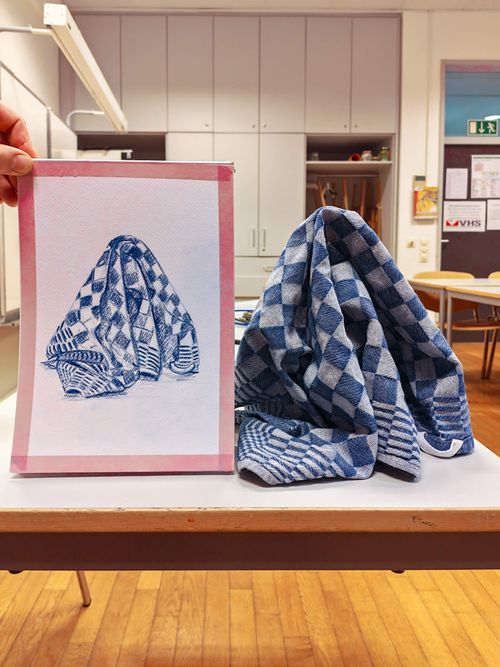 Student holds up a finished drawing of a folded blue-and-white checkered kitchen towel next to the actual towel